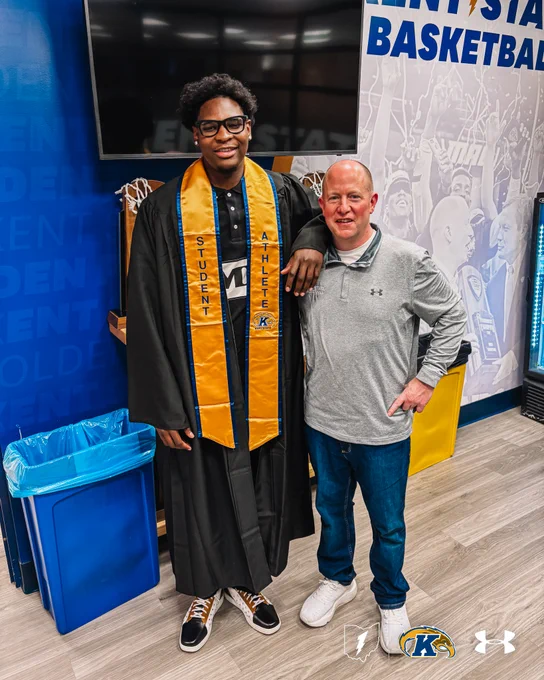 Delrecco Gillespie wearing graduation attire stands next to Head Coach Rob Senderoff dressed casually. They are the KSU MBB offices with Kent State University branding visible on the walls. The person in graduation attire is wearing a sash labeled "Student Athlete." The room has light wood flooring and a recycling bin. The Under Armour and Kent State logos are on the floor.