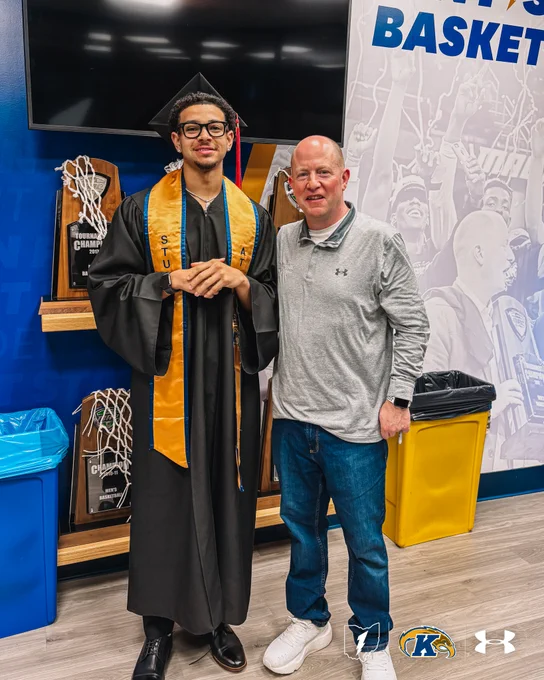 Brenden Moss in a cap and gown stands next to Head Coach Rob Senderoff in a casual outfit. They are in the KSU MBB offices with Kent State University basketball decor, including several trophies. Both individuals are smiling at the camera.