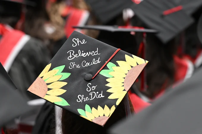 Close-up of a graduation cap worn in a seated crowd at a commencement ceremony. The black mortarboard is hand-painted with two large yellow sunflowers on opposite corners, green leaves extending inward, and a red tassel draped across the surface. In white script lettering, the cap reads, “She Believed She Could So She Did.” Other graduates wearing black caps with red accents are visible out of focus in the background.
