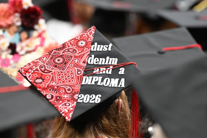 A decorated black graduation cap viewed from behind during a graduation ceremony. The left half of the cap is covered in red bandana-patterned fabric, while the right half remains black. Raised white letters spell out “dust and a DIPLOMA” with “2026” at the bottom. A red tassel lies across the cap. Rows of graduates in black caps and gowns fill the blurred background.