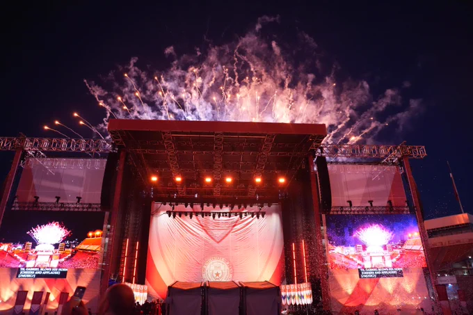 Fireworks explode above a stage featuring a white banner with 'THE UNIVERSITY OF TEXAS' seal. Screens display fireworks and text: 'TIL GABRIEL BLOWS HIS HORN [CHEERS AND APPLAUSE] [MUSIC]'. Stage lights illuminate the scene.