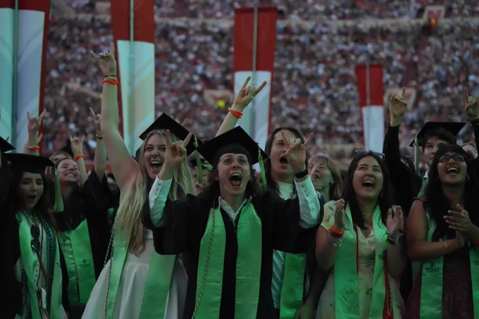 Graduates in caps and gowns cheer wildly at their commencement. A crowd fills the stadium behind them, with red and white banners visible. Many grads wear stoles marked \"TEXAS.\"