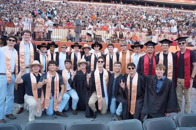 A crowd watches about twenty young men in graduation gowns and cowboy hats. The young men wear stoles that say TEXAS. The background is filled with many people in stadium seating and banners say \"TEXAS@2026\".