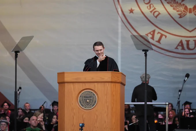 A man wearing graduation robes stands at a podium with \"THE UNIVERSITY OF TEXAS AT AUSTIN\" on it. He smiles and points at the viewer. Audience members and staff are visible in the background. A university logo is visible behind the speaker.