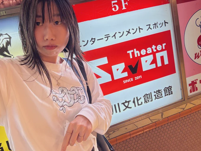 シアターセブンの看板の前で自撮りする人物(いといけいと)
着用した白い長袖Tシャツにはヘビのように身体の長いイルカが3匹絡み合ったイラストが刺繍されている
星のような形のシルバーのネックレスをつけており、肩くらいまで伸びた髪の毛先は外に向かって跳ねている