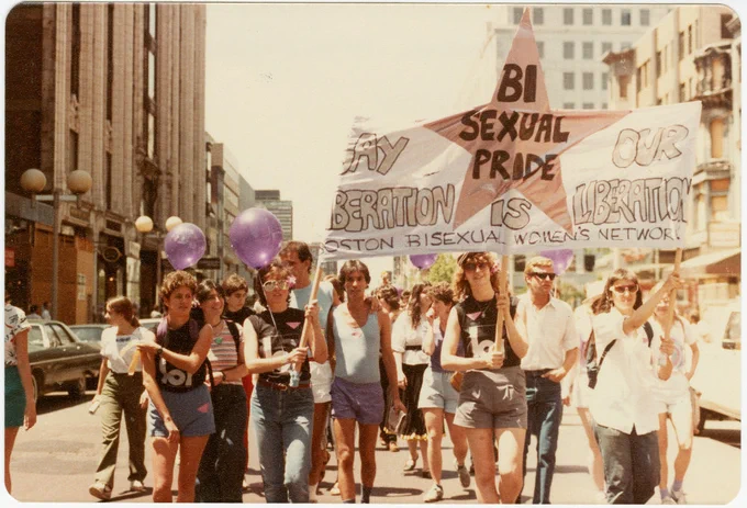 a protest sign being held up by a few people leading the crowd. the sign reads “GAY LIBERATION IS OUR LIBERATION. BISEXUAL PRIDE. BOSTON BISEXUAL WOMEN’S NETWORK”