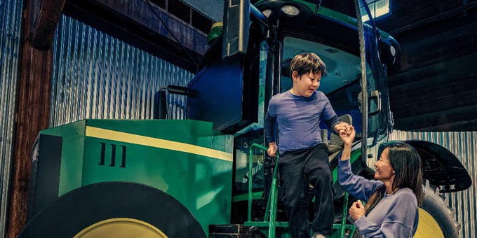 Child climbing down steps of a large green tractor inside a barn, helped by an adult woman holding his hand.