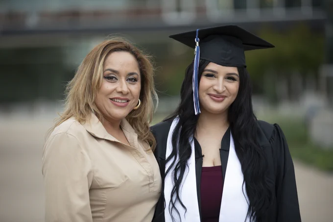 This image shows two individuals smiling outdoors. One person is wearing a graduation cap and gown with a white sash, suggesting a graduation event. The background features a blurred view of a building and greenery.