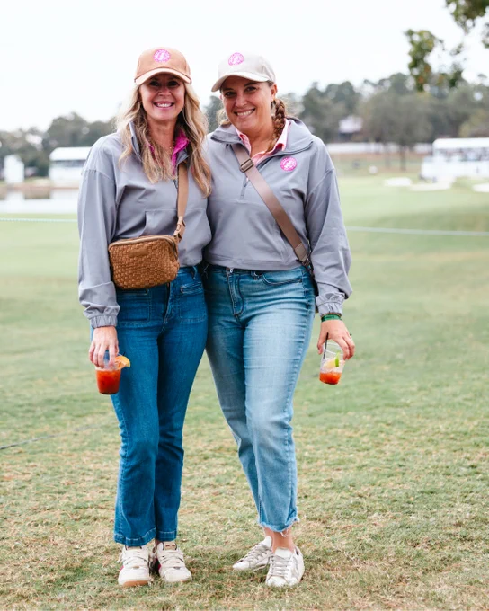 Two people standing on a grassy area, both wearing casual outfits including jeans, gray jackets, and caps with a logo. They are holding drinks garnished with fruit. Trees and a golf course can be seen in the background.