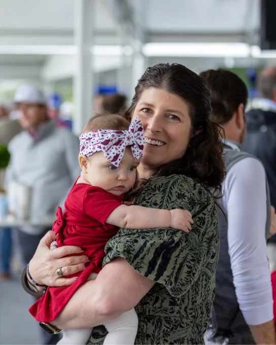 A person is smiling while holding a baby wearing a red outfit and a bow headband. Other people are in the background at a gathering.