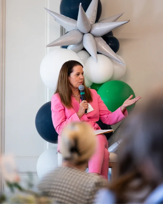 A person in a bright pink suit speaks into a microphone while seated on a chair. Behind is a decorative arrangement of black, white, green, and gray balloons. Audience members are visible in the foreground.