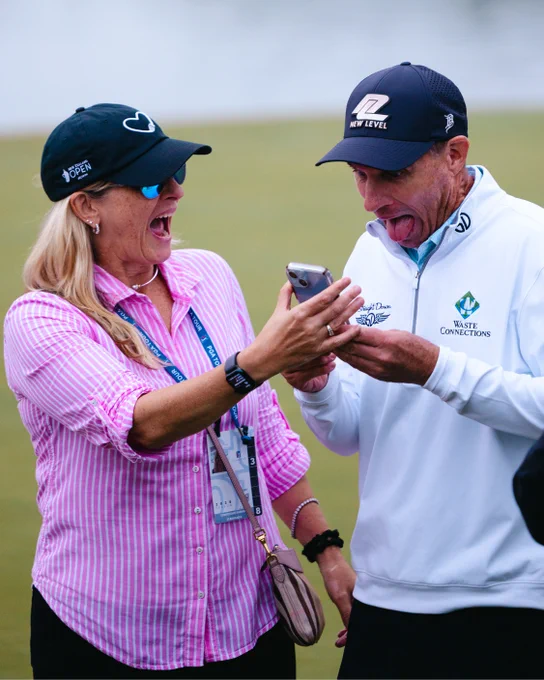 Two individuals are joyfully reacting while looking at a smartphone screen. They are standing on a golf course.