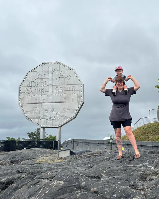 A mother smiling while giving her young child a shoulder ride in front of the iconic Big Nickel monument at Dynamic Earth in Sudbury.