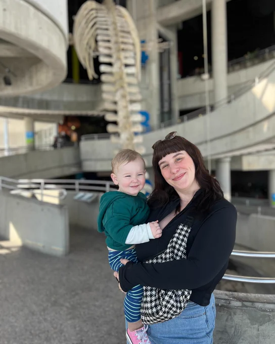 A mother holding her smiling toddler in the center of Science North, with the large Fin Whale skeleton exhibit visible in the background