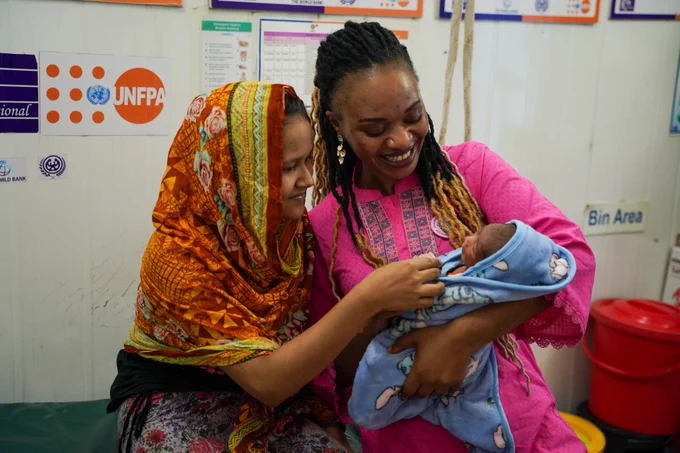 A photo of a mother, wearing a bright orange and yellow head scarf, and a midwife, in a bright pink tunic, smiling at a newborn baby wrapped in a blue blanket. The UNFPA logo is visible in the background.