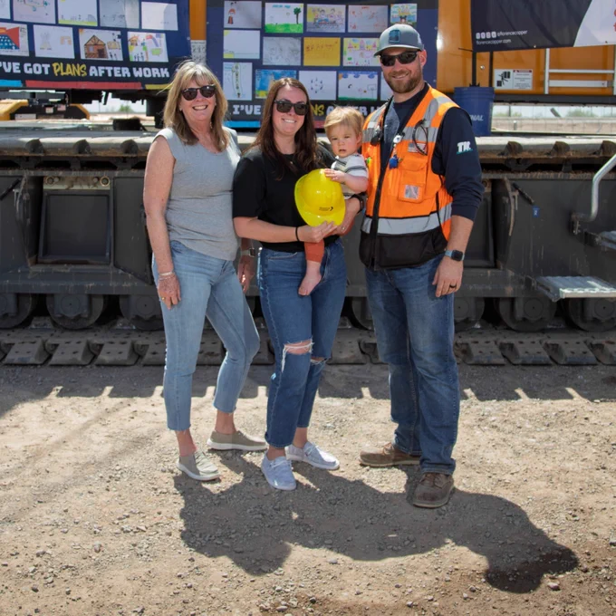 Four individuals are standing in front of a large construction vehicle, which has a display with various signs. One person is holding a child with a bright yellow helmet. Another person is wearing an orange safety vest and hat. The group is on a rocky surface, and part of the vehicle's treads are visible.
