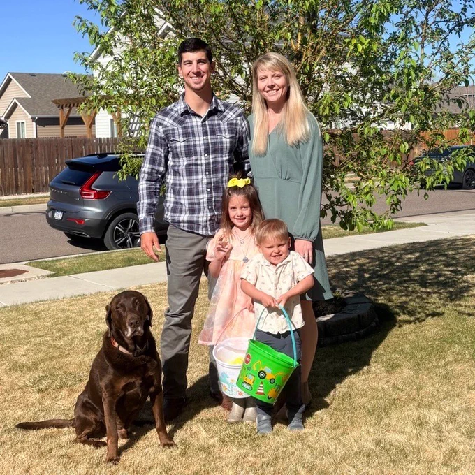 A family with two adults and two children stands in a yard on a sunny day. The children are holding a colorful bucket, and a brown dog sits nearby. There's a tree in the background, with houses and cars visible along the street.