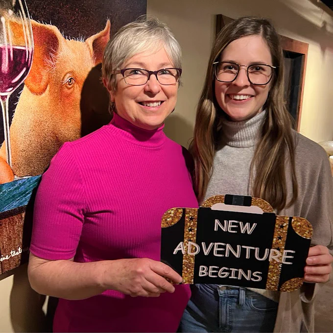 Two people are smiling and holding a sign that reads "New Adventure Begins." Behind them is a painting of a pig holding a glass of wine.