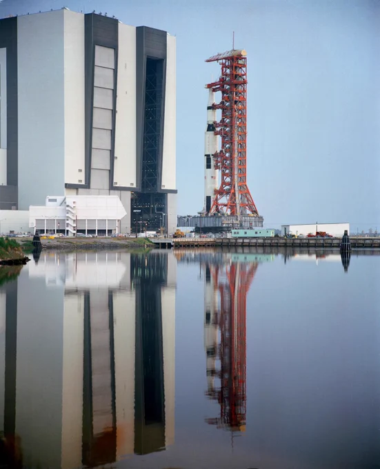 In this May 11, 1971, photo of the Apollo 15 Saturn V leaving the Vehicle Assembly Building (VAB), the rocket on the bright red mobile launcher and the VAB are perfectly reflected in a lagoon in the foreground.