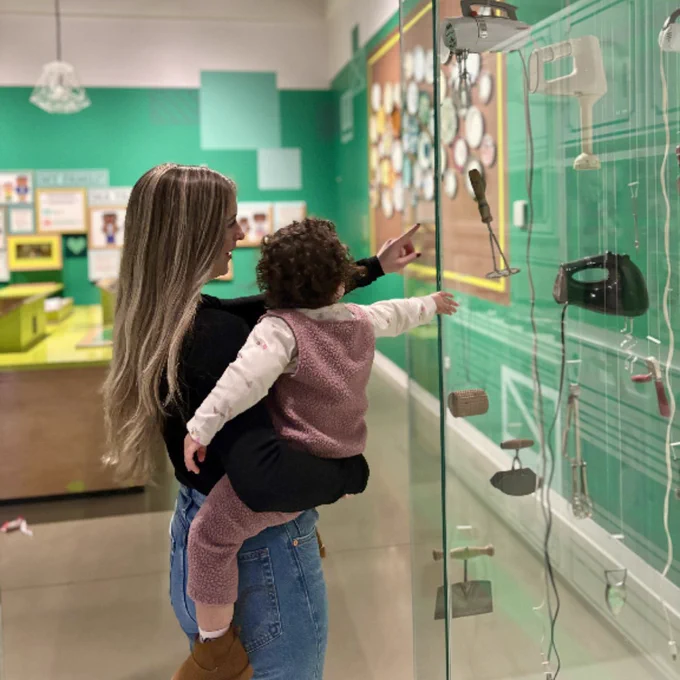 Une femme tenant un enfant dans ses bras montre du doigt des téléphones anciens exposés dans une vitrine lors d'une exposition dans un musée.