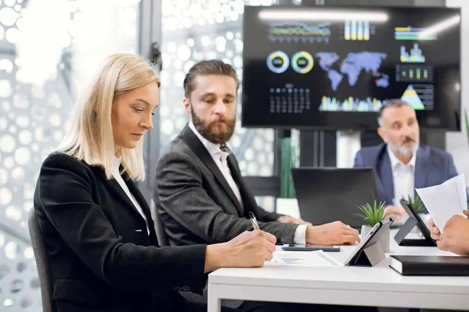 A group of individuals is seated at a conference table in a modern office, engaged in a meeting. A large screen displays charts and graphs in the background.
