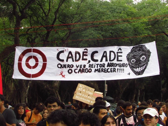 Faixa com a frase "Cadê, cadê, quero ver reitor arrombado o cargo merecer", com uma pintura de Aluísio Segurado, Reitor da Universidade de São Paulo (USP), com um olhar macabro. Créditos: Gabriel Albuquerque