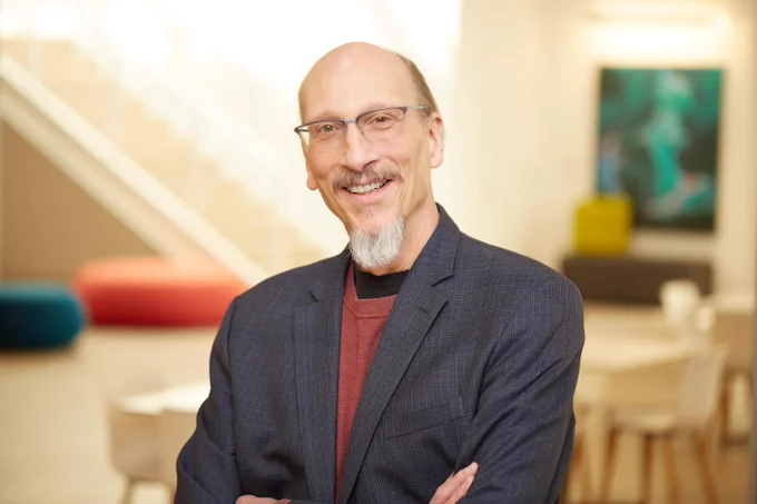 Disney Producer and Academy Award Winner Roy Conli stands indoors with arms crossed, shown from the chest up. They wear a dark checked blazer over a rust-colored sweater and a black undershirt. Conli has a short white goatee. The softly lit background shows a modern interior with colorful round seating, a staircase, a table and chairs, and a framed abstract artwork on the wall.