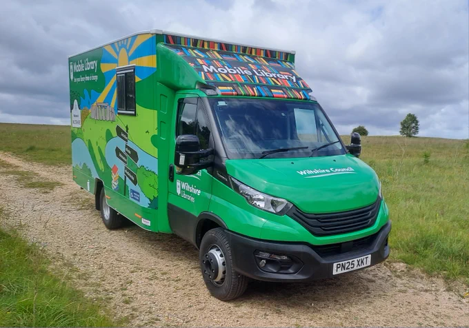 A Wiltshire Libraries green mobile van decorated in colourful book montage parked on a rural path against a countryside backdrop of green grass and trees in the distance.