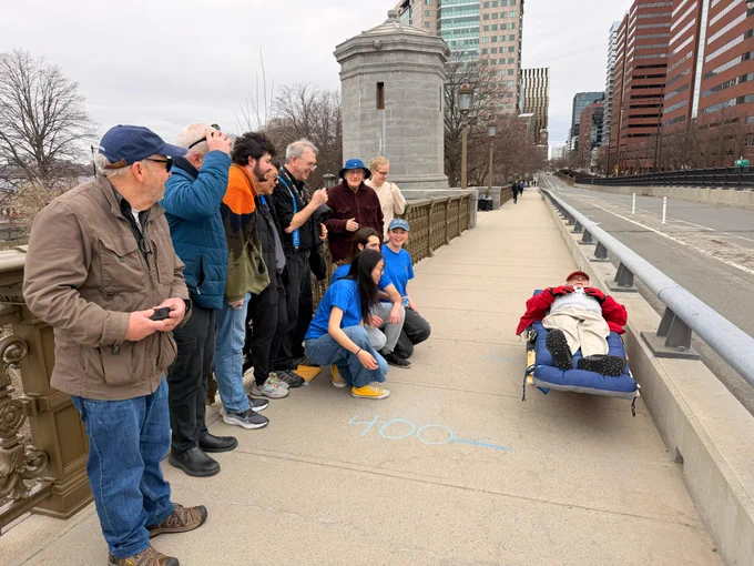Ten people stand and kneel on a bridge sidewalk next to a man lying on a wooden cart.