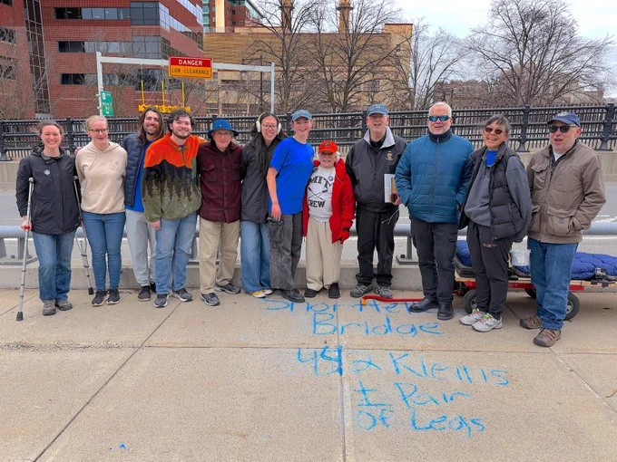 A group of 12 people stand together on a bridge with the writing, “Shortfellow Bridge - 442 Kleins   pair of legs” in blue chalk on the sidewalk.