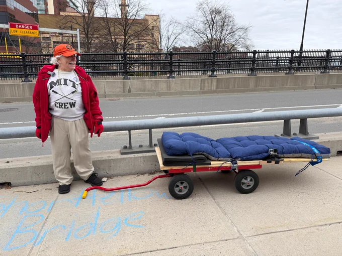 A man in an MIT sweatshirt, red coat, and gloves stands next to a wooden cart on a bridge.