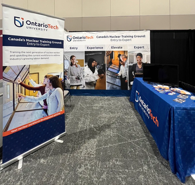 A conference booth for Ontario Tech University set up against a neutral wall. On the left, a vertical banner reads “Canada’s Nuclear Training Ground: Entry to Expert” and highlights training for the next generation of nuclear workers, with an image of people interacting with a large touchscreen display. In the center, a wide backdrop displays four stages—Entry, Experience, Elevate, and Expert—each paired with photos of students or professionals engaged in hands-on learning and technical work. On the right, a table covered with an Ontario Tech University tablecloth holds two monitors and neatly arranged promotional materials. The floor is carpeted, and the booth appears ready for attendees but is currently unoccupied.