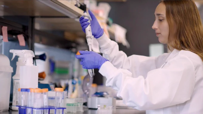 A person in a lab coat and gloves uses a pipette in a laboratory setting. Shelves with various lab equipment and supplies are visible in the background.