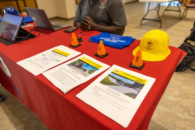 Table with attendee waiting. On table are flyers about construction projects and giveaways including safety cone stress toys, plastic construction hats, and cooling towels.