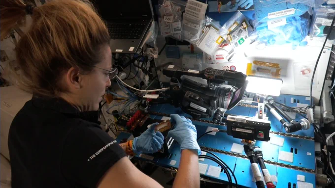 ESA Astronaut Sophie Adenot works on CBIOMES aboard the space station. Her workstation is set up above several metallic blue panels. She holds a rectangular compartment that will be photographed for the CBIOMES experiment.