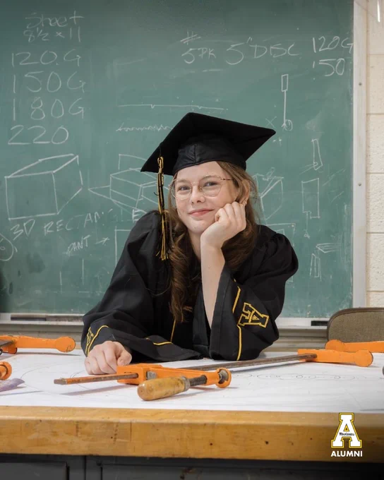Zoe Moore poses in her cap and gown at a drafting table in a woodworking classroom, with tools and design sketches visible on the chalkboard behind her.