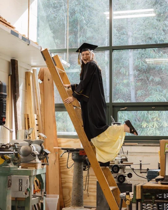 Zoe Moore smiles while climbing a wooden ladder in her cap and gown inside a woodworking studio, surrounded by equipment and large workshop windows.