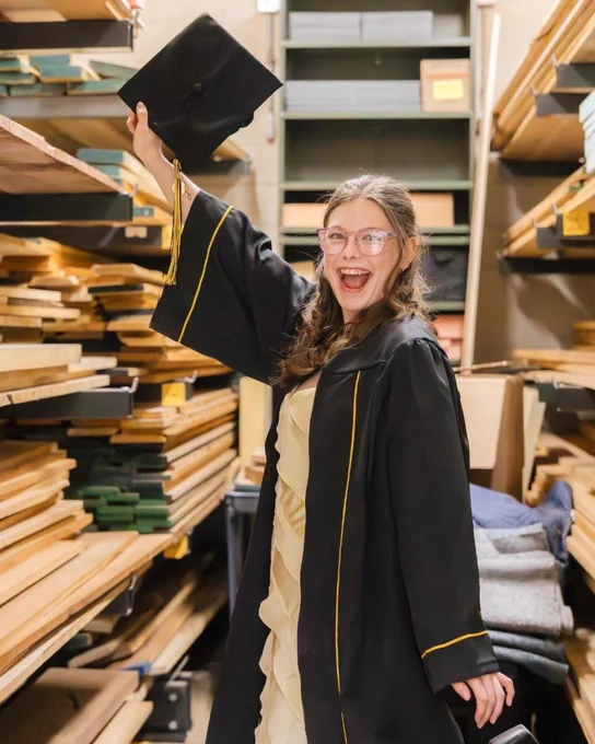 Zoe Moore celebrates in her cap and gown while holding her graduation cap in a woodshop storage area filled with stacked lumber.
