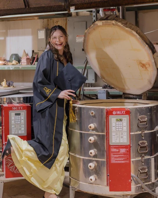 Zoe Moore laughs beside a large ceramics kiln in her cap and gown inside an art studio workspace.