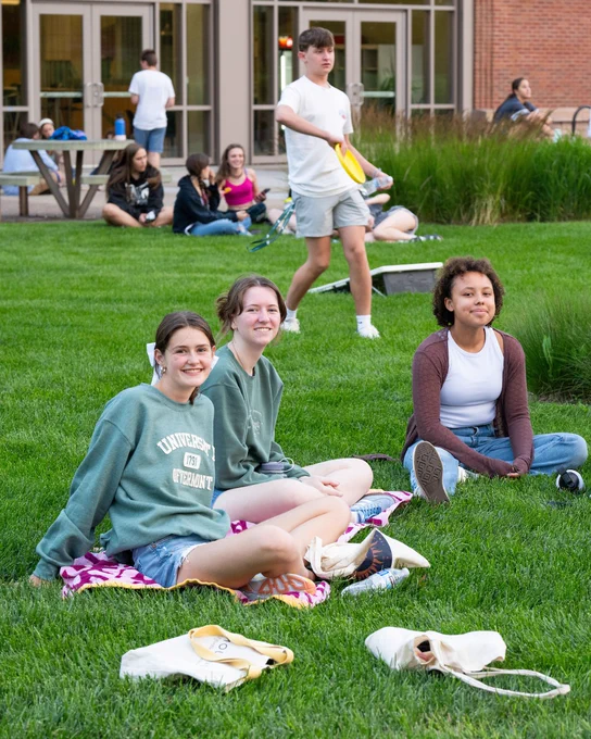 Three people are seated on a grassy lawn with University of Vermont logos on their clothing. They are smiling toward the camera. In the background, other individuals are engaged in outdoor activities such as playing catch with a frisbee. A building with large windows is visible behind them.