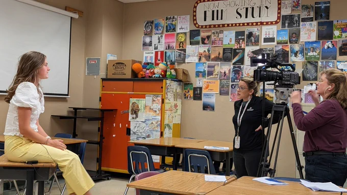 A female student is sitting on a desk that is facing two adults with a video camera on a tripod.