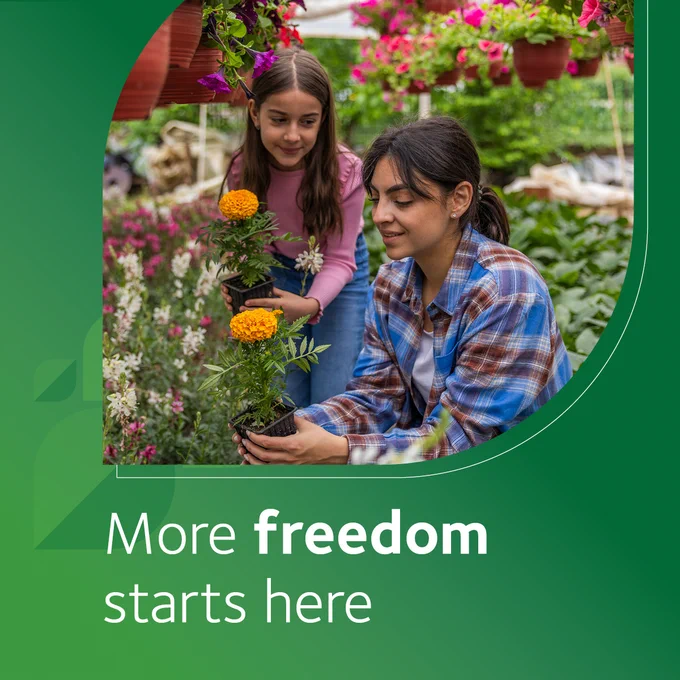 A woman and young girl gardening together, surrounded by a variety of potted flowers and hanging plants. They are handling a bright orange marigold. Text on the image reads, "More freedom starts here.