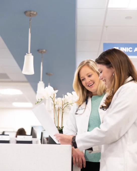 A Tryon Medical Partners doctor and advanced care clinician smile as they look together at a document standing at the nurses' station in a medical office