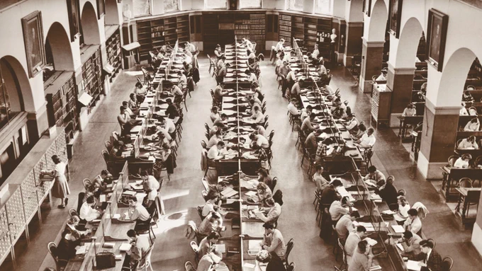 A sepia-toned image of a busy library reading room, filled with long tables and numerous individuals engaged in reading and writing. The room has tall arches, bookshelves lining the walls, and a high ceiling, indicating a grand and historic setting.