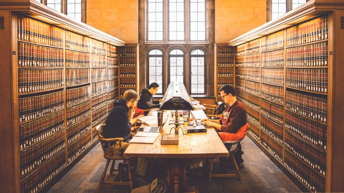 A group of people studying at a long wooden table in a library lined with shelves full of books. Large arched windows in the background let in natural light.