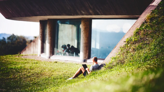 Side view of a modern concrete building with a large glass window. A person is sitting on a grassy slope in the foreground, using a laptop. The scene is peaceful and bathed in natural light.