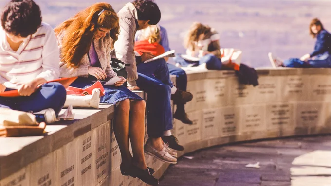 Students sit on a curved stone wall outdoors, studying and writing in notebooks.