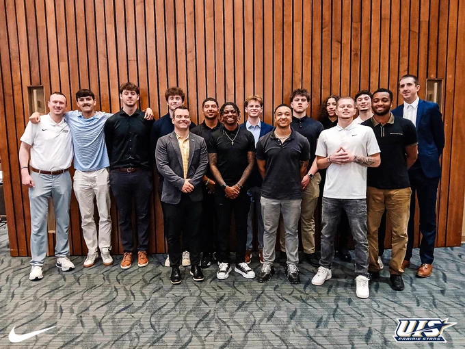The UIS men’s basketball team and coaching staff pose together in dress attire at the Night of the Stars banquet in front of a wood-paneled wall.