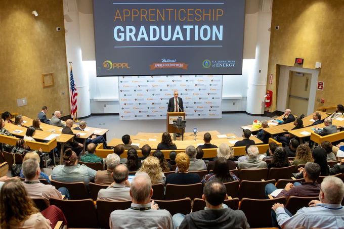 Princeton University's Peter Schieffer stands at a podium in a lecture hall during an Apprenticeship Graduation event. The backdrop displays logos for PPPL, National Apprenticeship Week, and the U.S. Department of Energy. The room is filled with seated attendees.