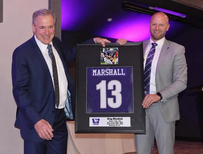 Greg Marshall and Michael Faulds hold up a jersey from of Marshall’s old number 13 jersey that includes a photo of Marshall playing for the Western Mustangs at the top of the frame.